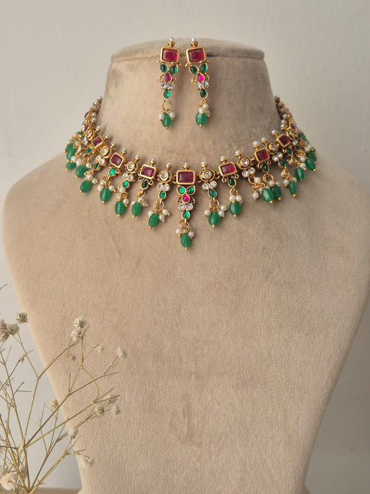 An ornate gold-plated choker featuring square ruby-red stones, emerald-green beads, and delicate pearl drops with matching earrings by naitri.in