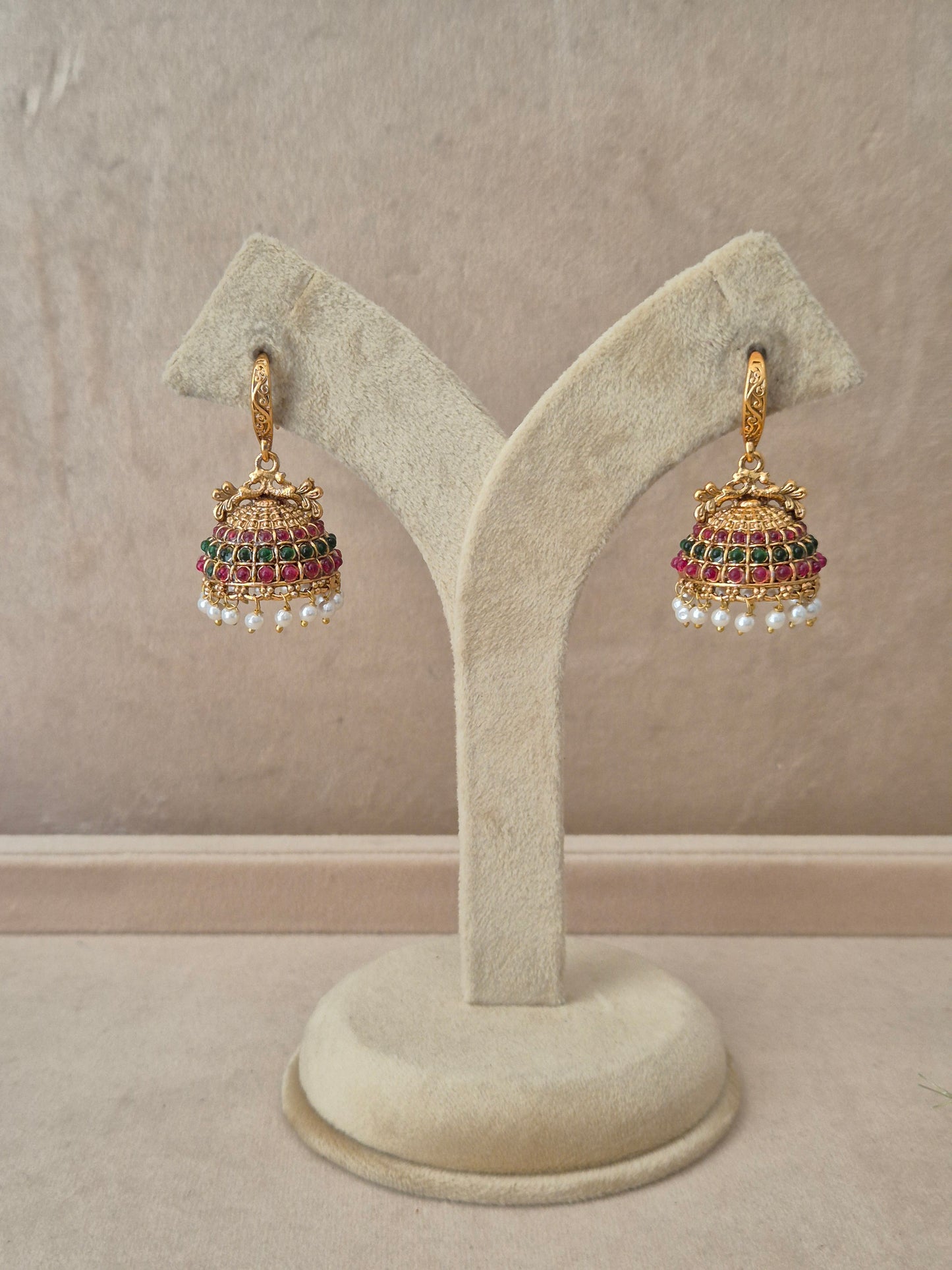 Pair of gold-plated Hemakshi Jhumka earrings hanging on a beige velvet display stand, featuring detailed textured hooks and traditional bell-shaped drops adorned with rows of red and green gemstones and small white dangling pearls by naitri.in