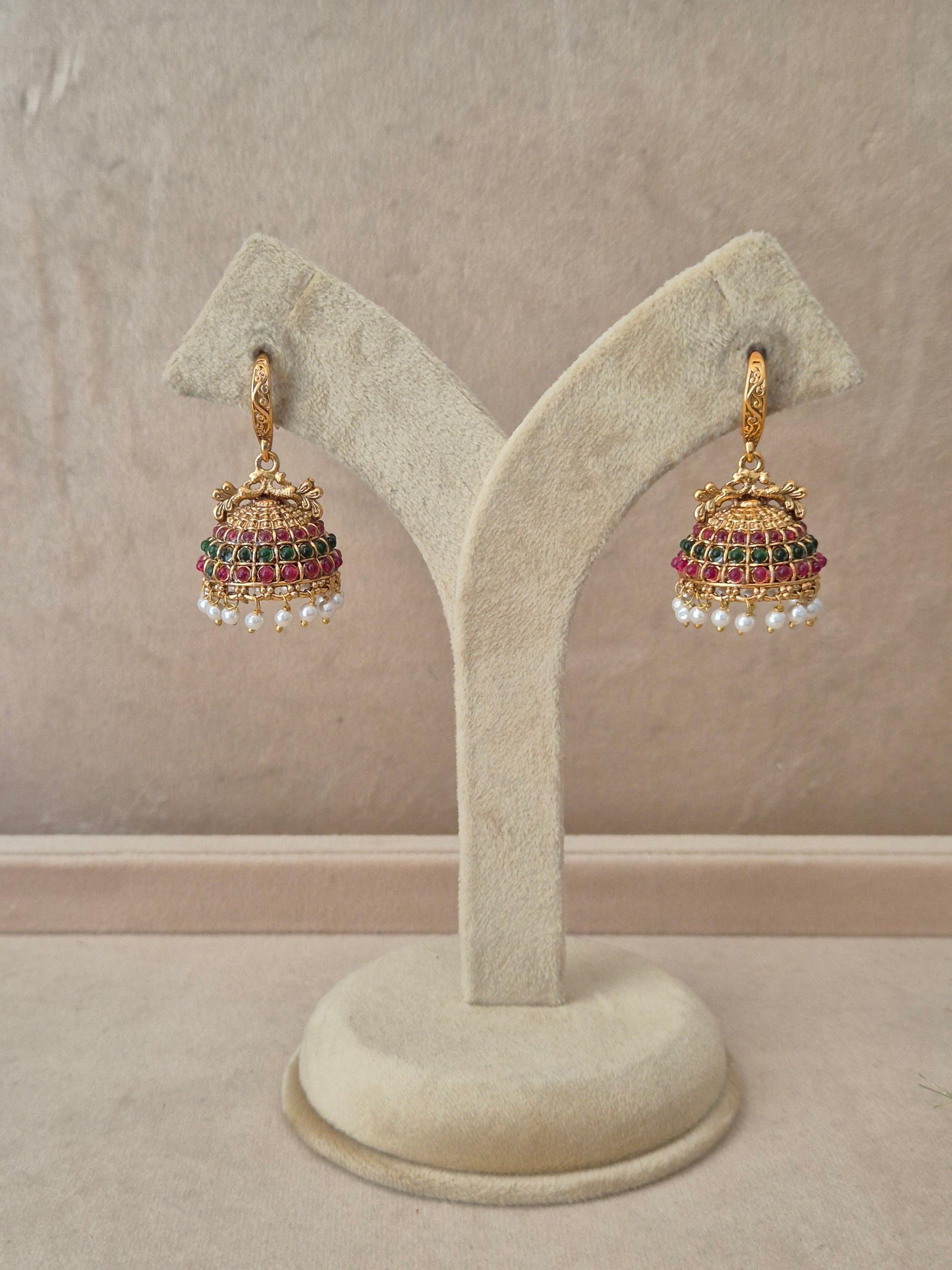 Pair of gold-plated Hemakshi Jhumka earrings hanging on a beige velvet display stand, featuring detailed textured hooks and traditional bell-shaped drops adorned with rows of red and green gemstones and small white dangling pearls by naitri.in