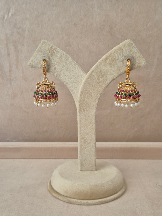 Pair of gold-plated Hemakshi Jhumka earrings hanging on a beige velvet display stand, featuring detailed textured hooks and traditional bell-shaped drops adorned with rows of red and green gemstones and small white dangling pearls by naitri.in