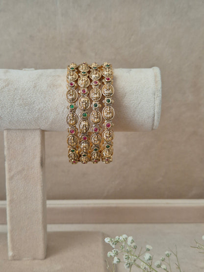 A set of four gold-plated Hemali bangles featuring traditional Indian temple jewelry engraving with alternating red and green gemstone accents, displayed on a beige fabric jewelry stand by naitri.in