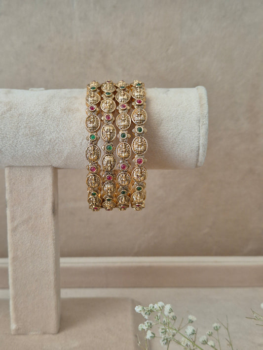 A set of four gold-plated Hemali bangles featuring traditional Indian temple jewelry engraving with alternating red and green gemstone accents, displayed on a beige fabric jewelry stand by naitri.in