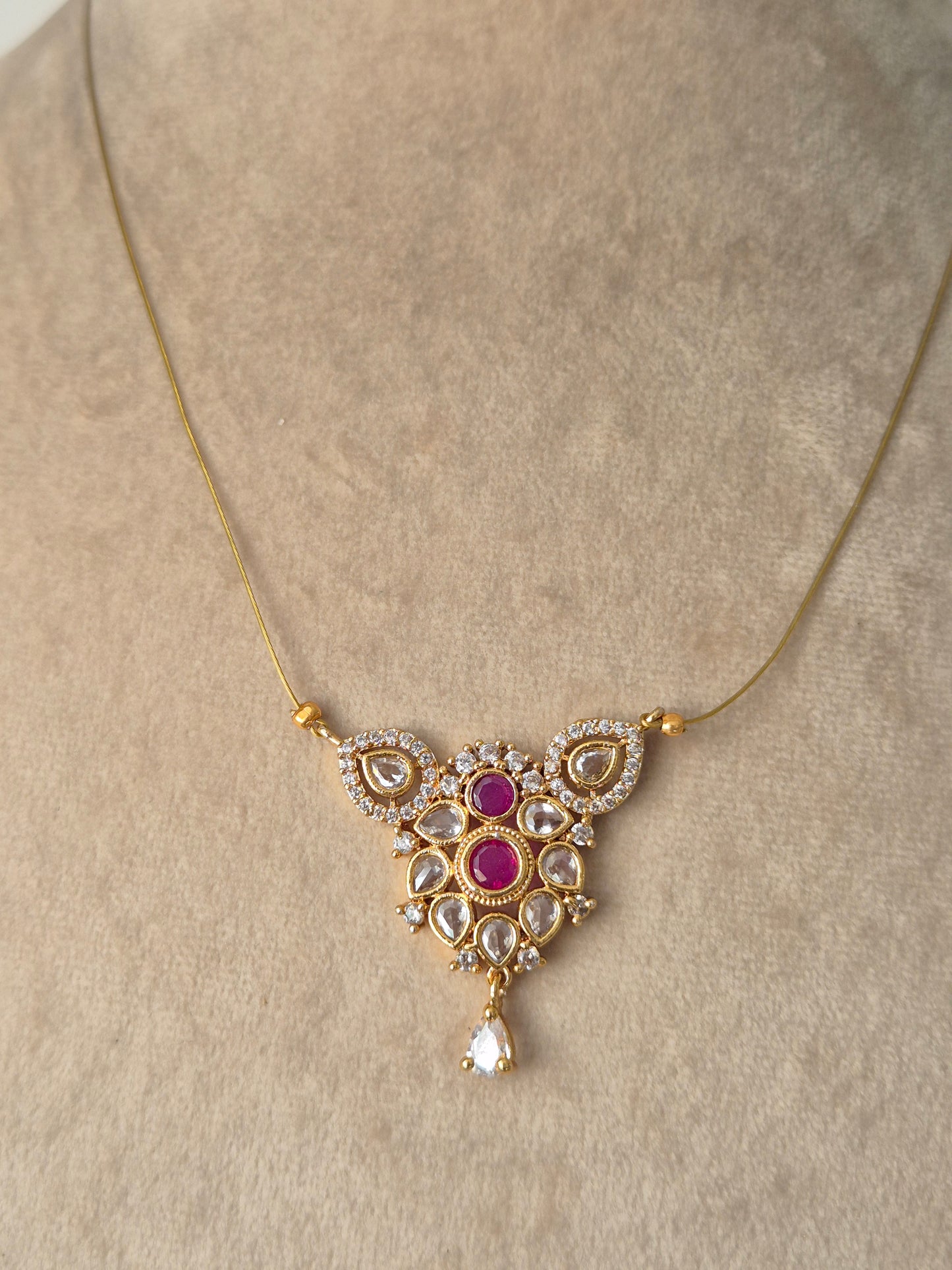 A delicate gold necklace  featuring an ornate central pendant with pink gemstones surrounded by small white stones in a floral and teardrop design, finished with a single dangling teardrop gem by naitri.in
