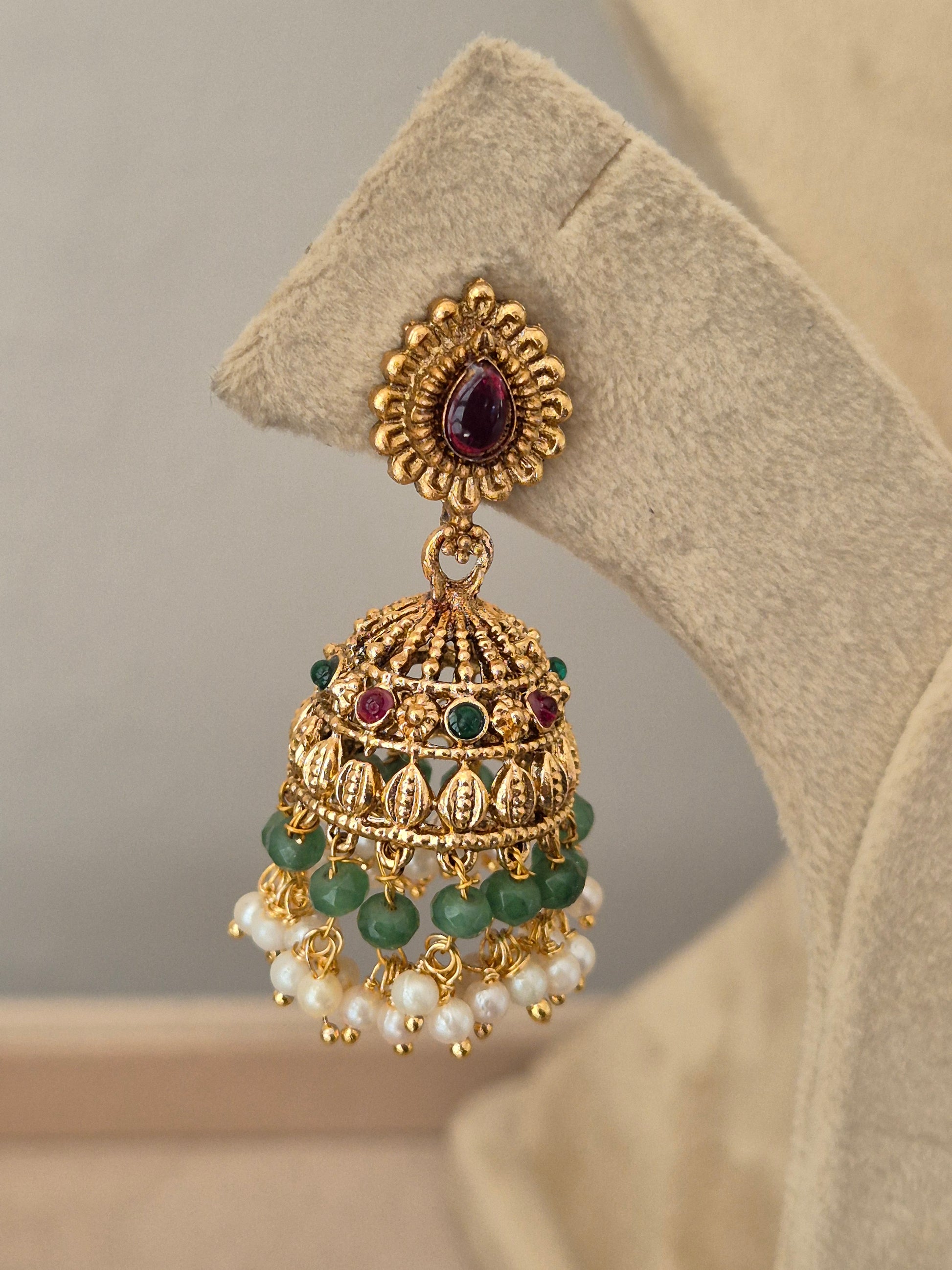 An ornate gold-toned traditional Indian jhumka earring the earring features a teardrop-shaped purple stone in the front, a detailed filigree dome with green stone accents, and dangling strands of white faux pearls by naitri.in
