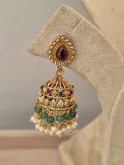 An ornate gold-toned traditional Indian jhumka earring the earring features a teardrop-shaped purple stone in the front, a detailed filigree dome with green stone accents, and dangling strands of white faux pearls by naitri.in