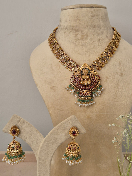 Gold-toned traditional Indian jewelry, including a necklace, earrings, and a matching stand, with a central ornate piece featuring gold, red, green, and white details by naitri.in