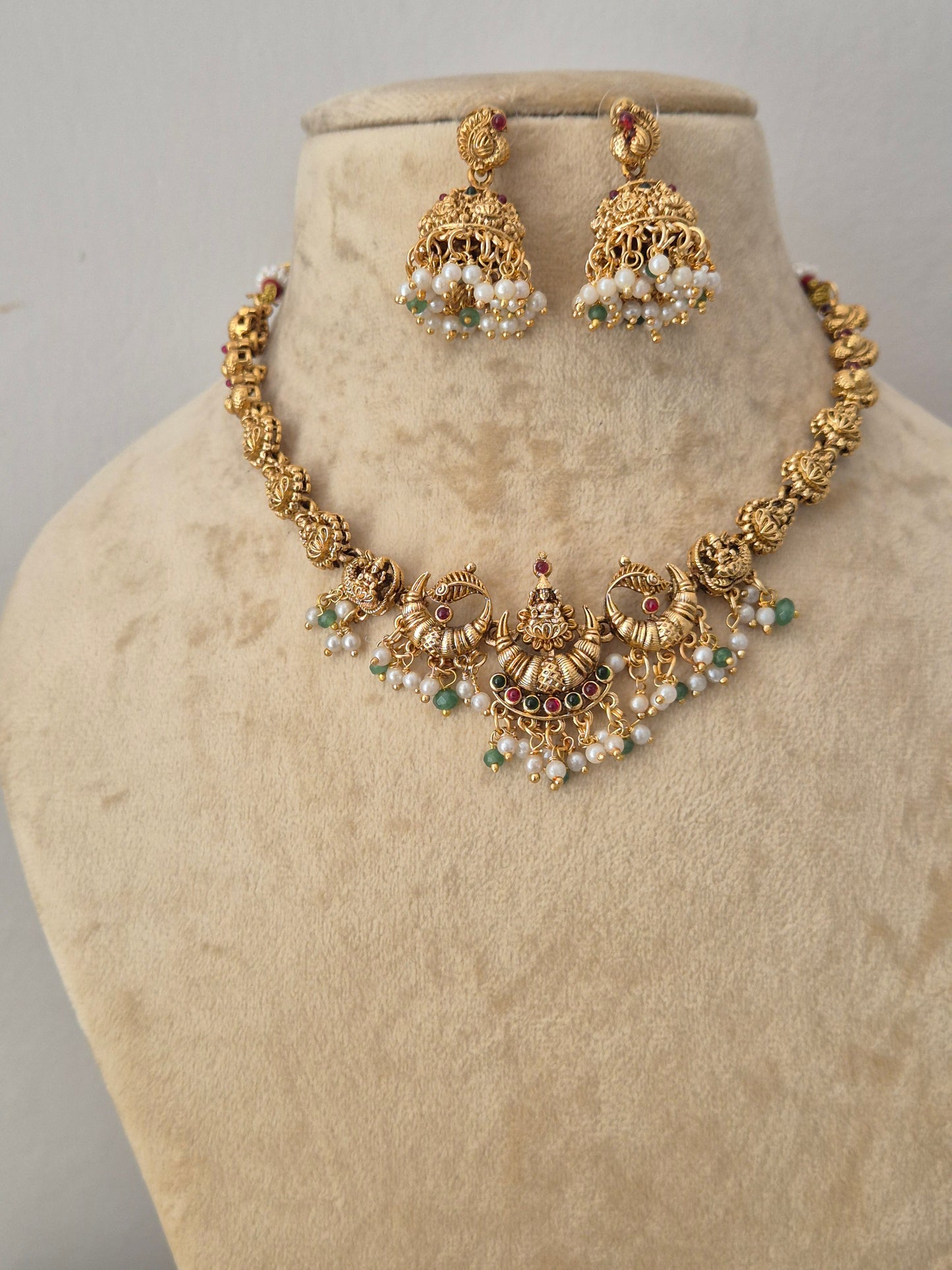 Gold-toned traditional Indian jewelry set featuring a necklace and matching earrings, the jewelry is decorated with gold filigree, white faux pearls, and small green and red stone accents by naitri.in