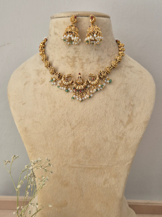 Gold-toned traditional Indian jewelry set featuring a necklace and matching earrings. The jewelry is decorated with gold filigree, white faux pearls, and small green stone accents by naitri.in