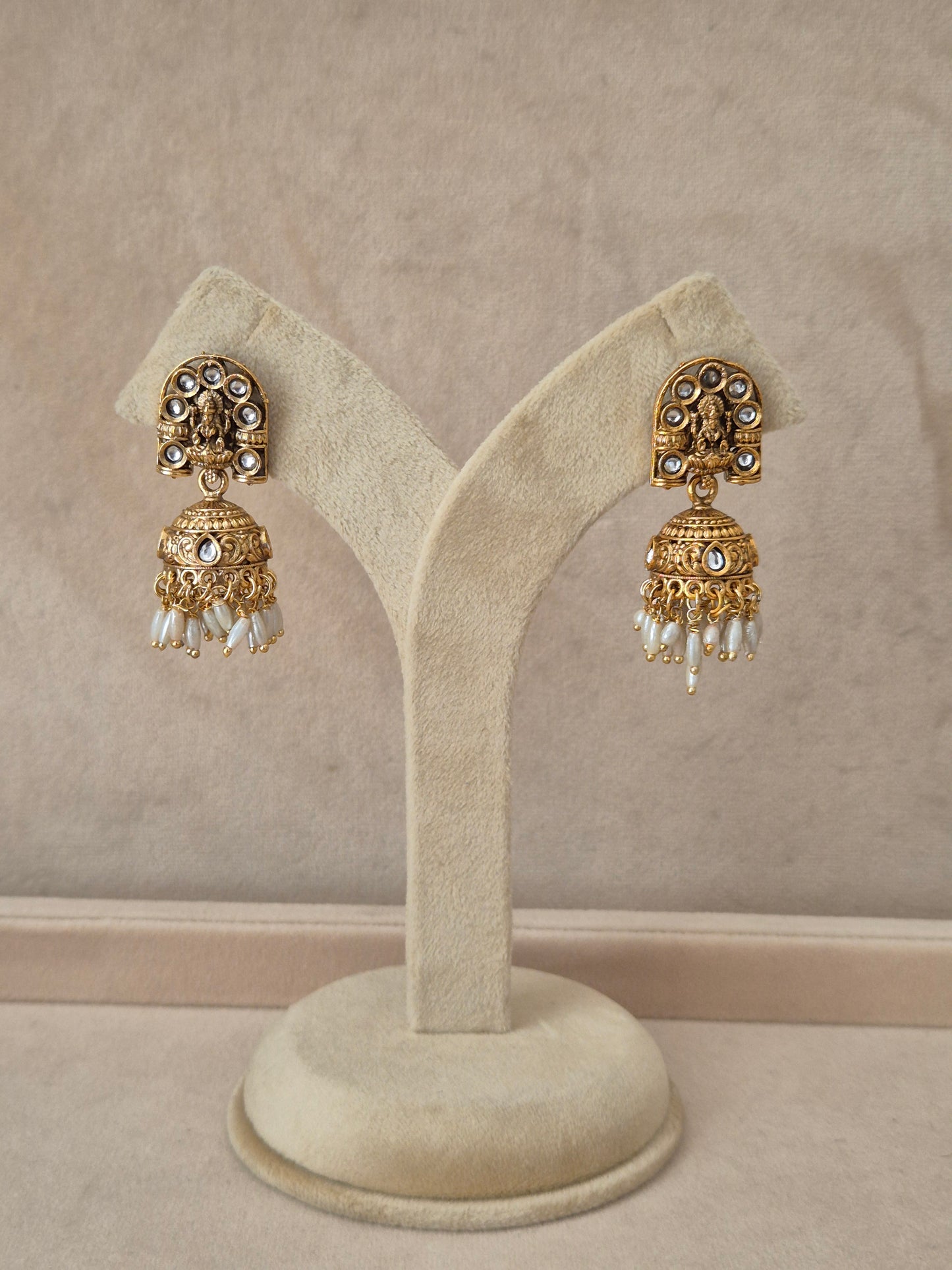 Pair of gold-toned Jaimini Jhumka earrings hanging on a beige velvet display stand, featuring traditional engraved studs with small white pearl drops. by naitri.in