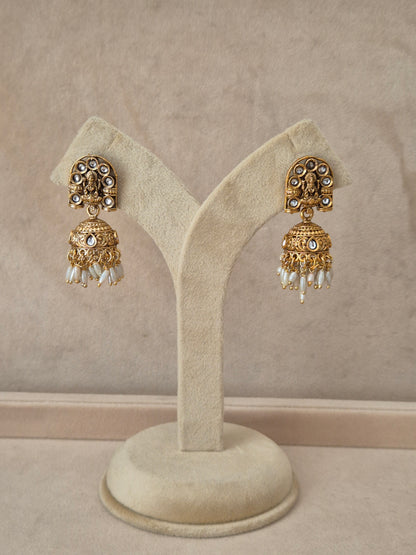 Pair of gold-toned Jaimini Jhumka earrings hanging on a beige velvet display stand, featuring traditional engraved studs with small white pearl drops. by naitri.in
