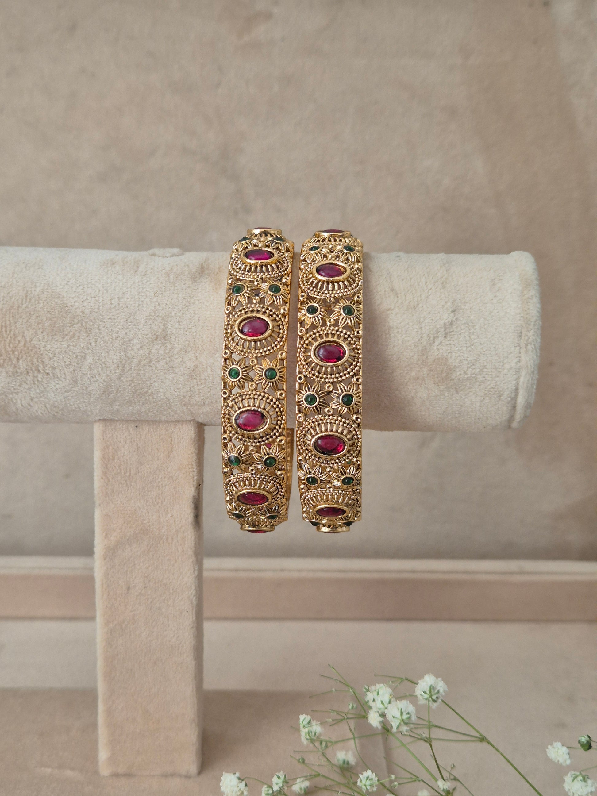 A pair of ornate gold-toned bangles with red and green stone accents, by naitri.in