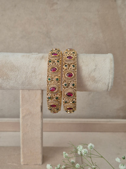 A pair of ornate gold-toned bangles with red and green stone accents, by naitri.in
