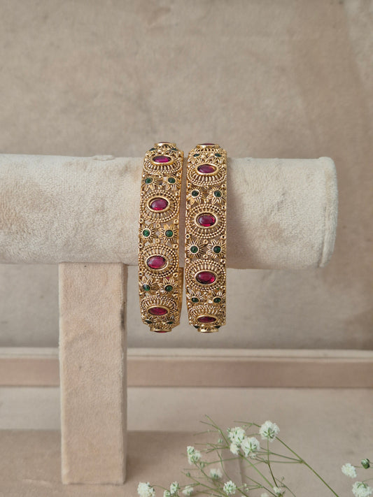 A pair of ornate gold-toned bangles with red and green stone accents, by naitri.in