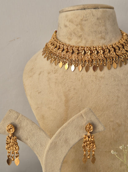 Gold-toned traditional Indian choker necklace and matching long dangle earrings set. The choker features detailed textured metalwork of leaf and floral motifs with dangling flat, teardrop-shaped metallic charms, paired with coordinating drop earrings featuring intricate filigree by naitri.in