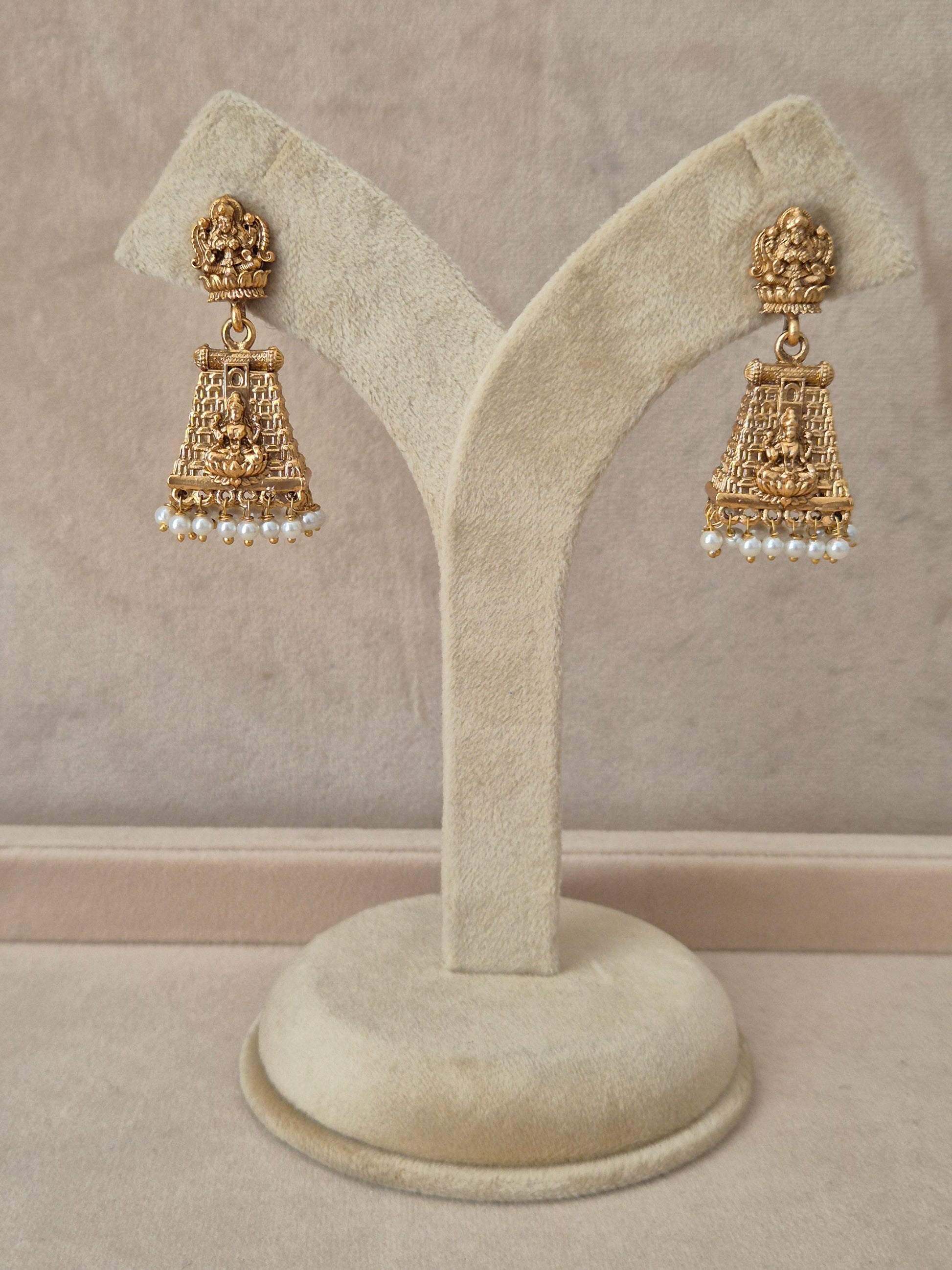 Pair of gold-toned Jayashree temple jewelry earrings hanging on a beige velvet display stand, featuring traditional engraved drops lined with small white pearls at the bottom by naitri.in