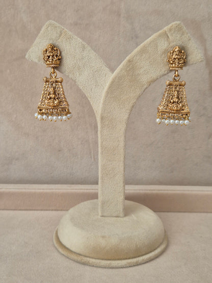 Pair of gold-toned Jayashree temple jewelry earrings hanging on a beige velvet display stand, featuring traditional engraved drops lined with small white pearls at the bottom by naitri.in