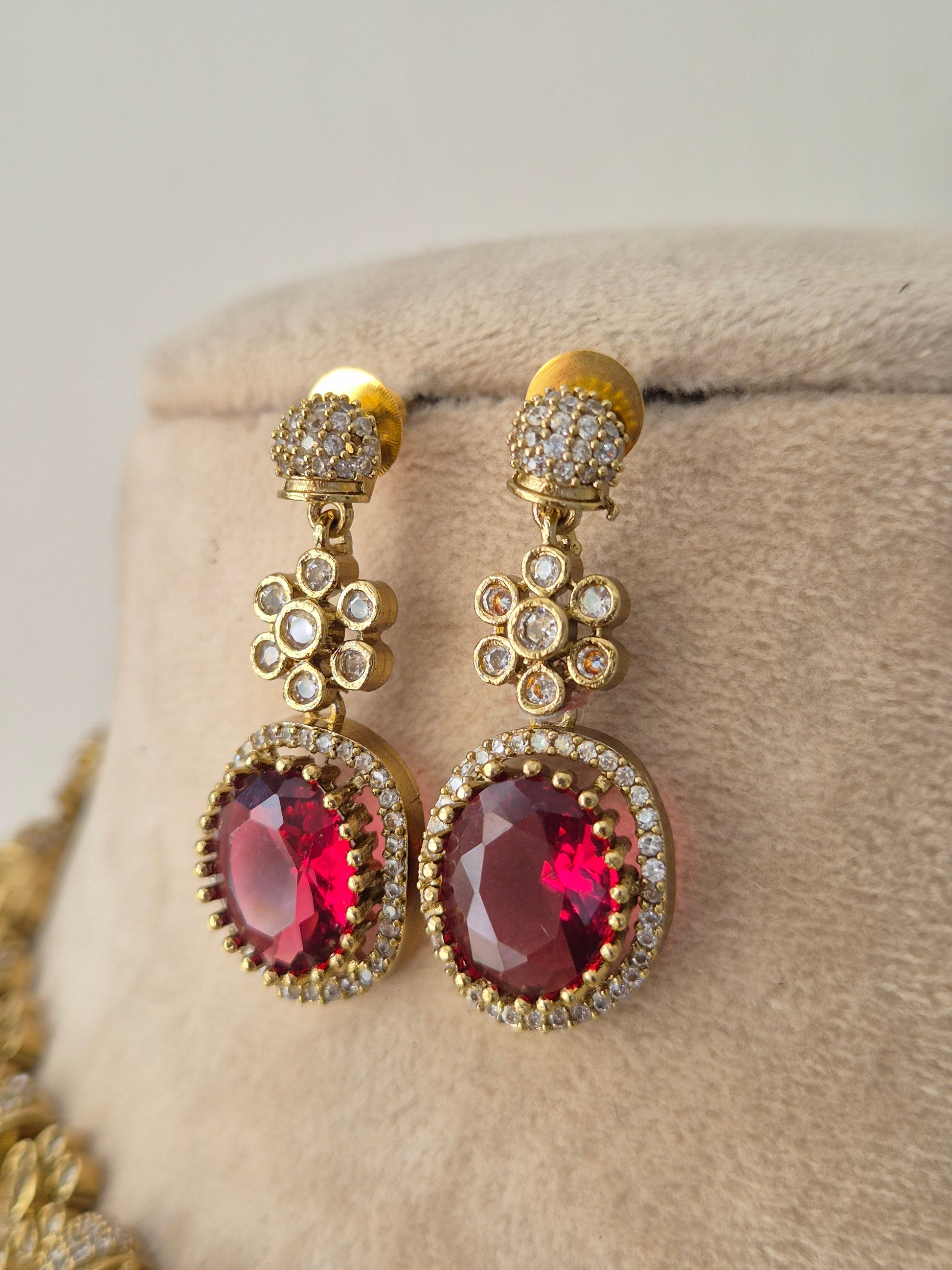 A pair of gold-toned drop earrings with oval pinkish-red gemstones surrounded by small clear stones, by naitri.in
