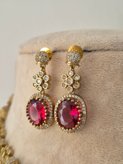 A pair of gold-toned drop earrings with oval pinkish-red gemstones surrounded by small clear stones, by naitri.in