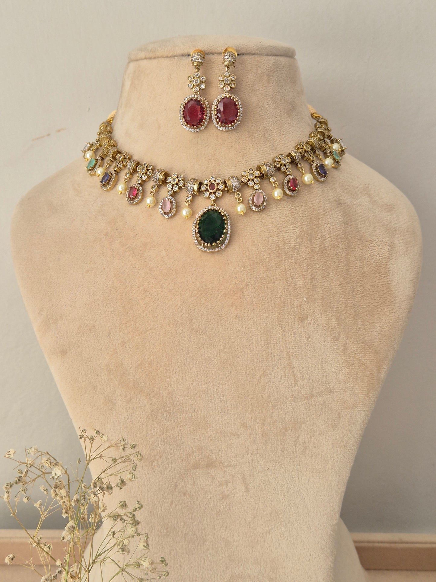 Gold-toned traditional Indian jewelry set featuring a necklace and matching earrings, The necklace has a gold chain decorated with small white pearls, a central oval green stone, and multiple round stones in red, pink, and blue, along with gold floral motifs naitri.in