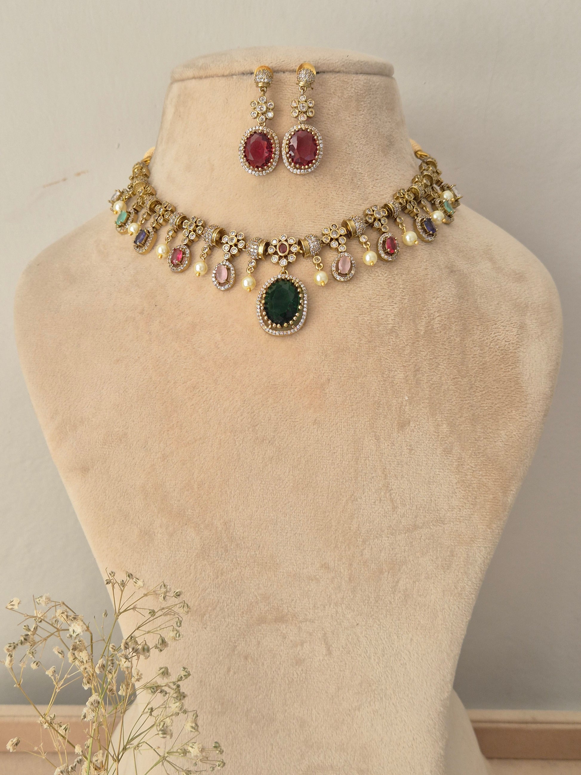 Gold-toned traditional Indian jewelry set featuring a necklace and matching earrings, The necklace has a gold chain decorated with small white pearls, a central oval green stone, and multiple round stones in red, pink, and blue, along with gold floral motifs naitri.in