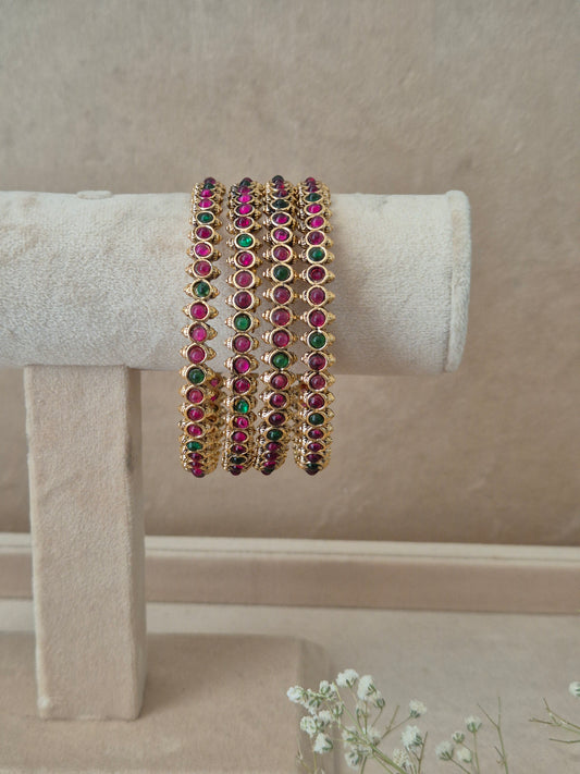 A set of four gold-plated Kadambari bangles lined up showcasing a dense pattern of small, round, bezel-set red and green gemstones with tiny gold bead borders. by naitri.in