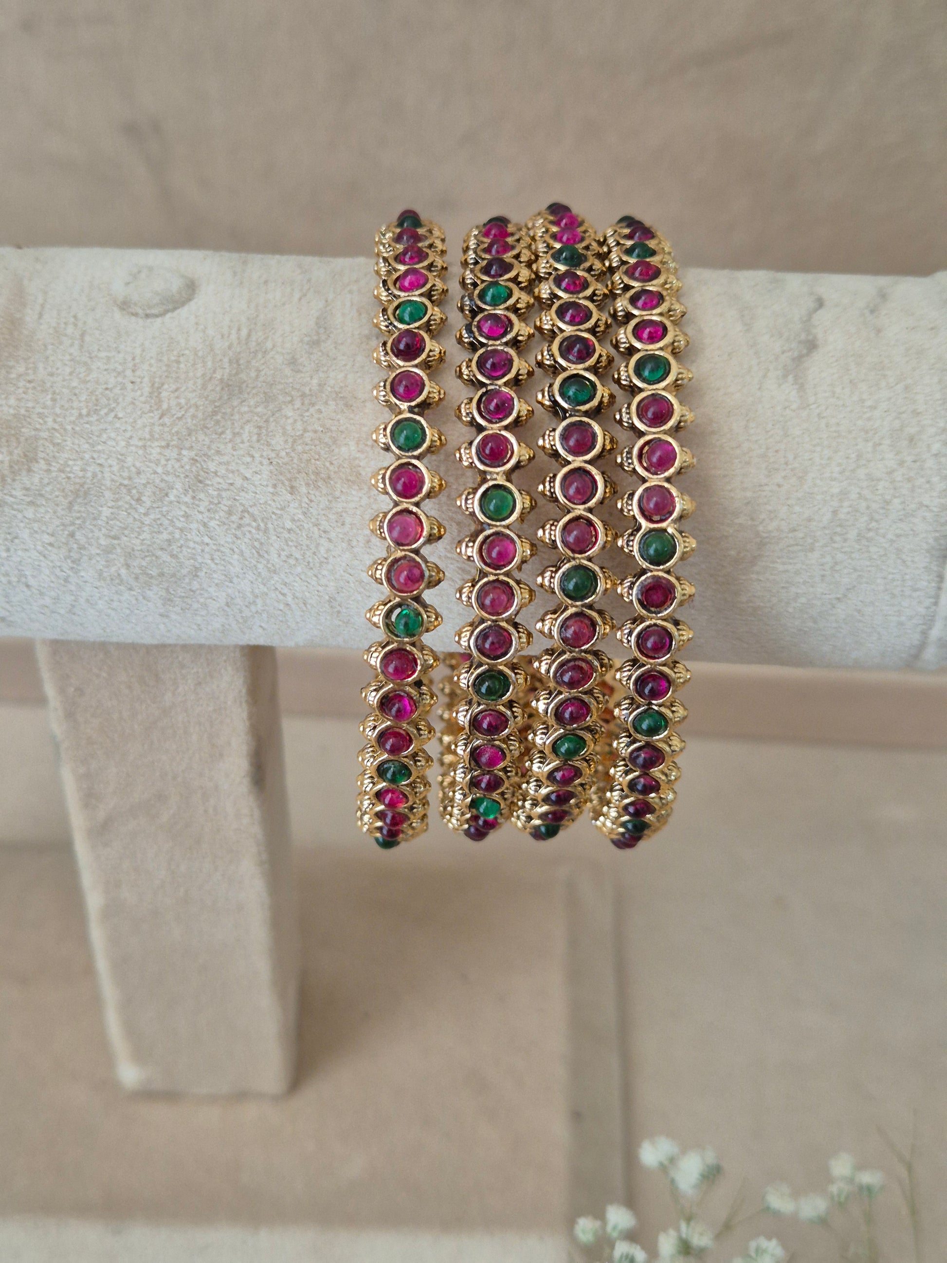 Four traditional golden festive bangles decorated with circular rows of alternating red and green stone settings flanked by intricate gold granulated beadwork details. By Naitri.in