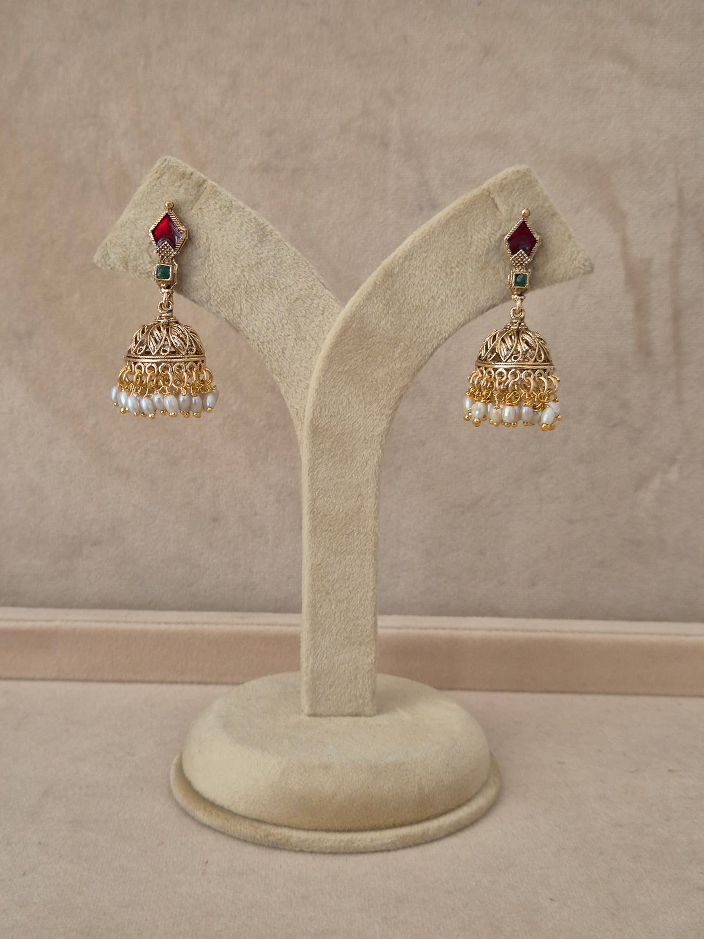 Pair of gold-toned Kalindi Jhumka earrings hanging on a beige velvet display stand, featuring diamond-shaped studs with red and green gemstones and detailed bell-shaped drops fringed with small white pearls by naitri.in