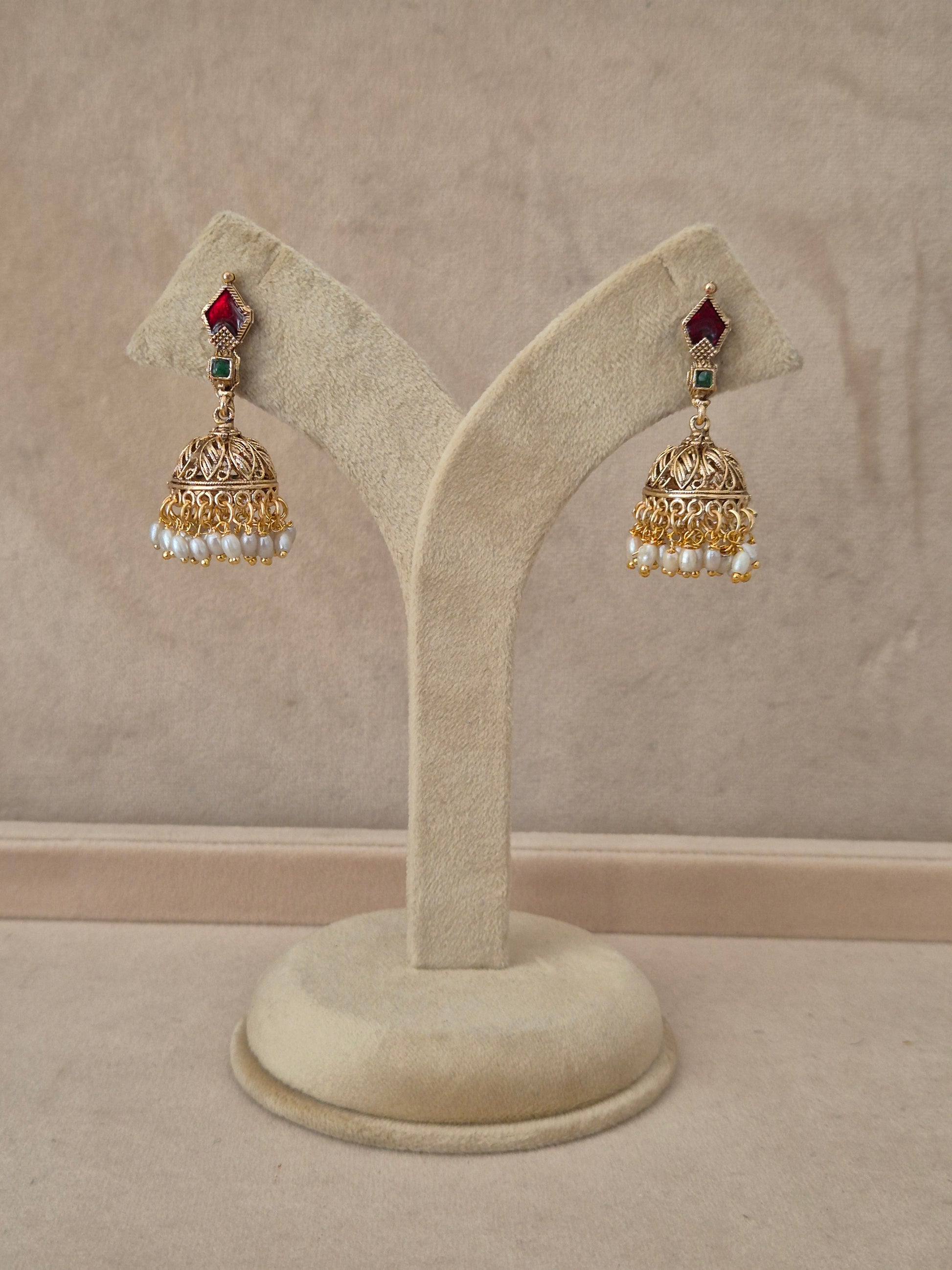 Pair of gold-toned Kalindi Jhumka earrings hanging on a beige velvet display stand, featuring diamond-shaped studs with red and green gemstones and detailed bell-shaped drops fringed with small white pearls by naitri.in