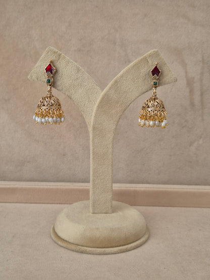 Pair of gold-toned Kalindi Jhumka earrings hanging on a beige velvet display stand, featuring diamond-shaped studs with red and green gemstones and detailed bell-shaped drops fringed with small white pearls by naitri.in