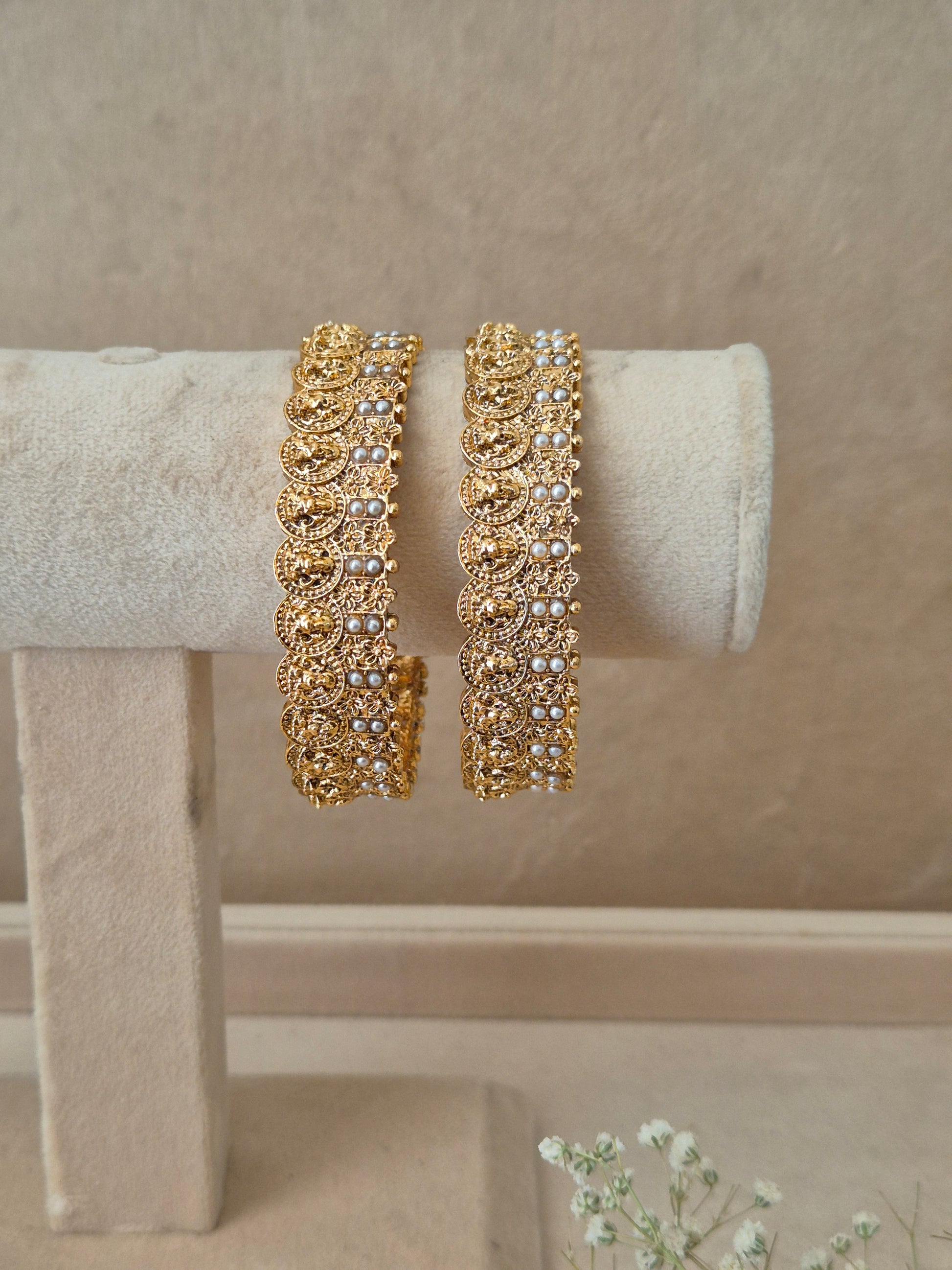 A pair of gold-plated Kanishka bangles featuring intricate traditional floral patterns and delicate white pearl accents along the edges, displayed on a beige velvet jewelry stand by naitri.in