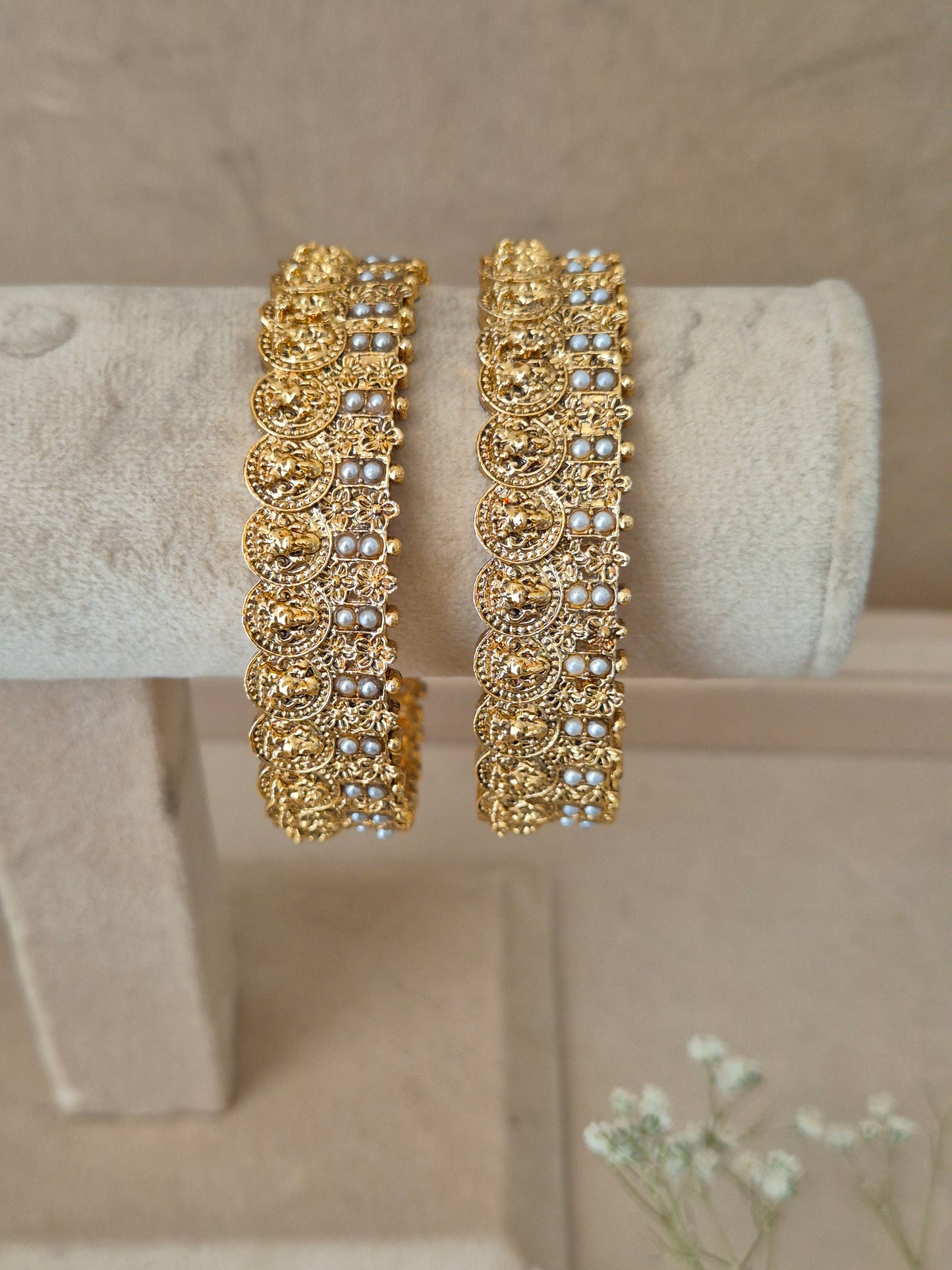 Elegant golden statement bangles with ethnic details exhibited on a neutral luxury jewelry display next to small white flowers by naitri.in