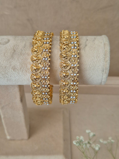 Elegant golden statement bangles with ethnic details exhibited on a neutral luxury jewelry display next to small white flowers by naitri.in