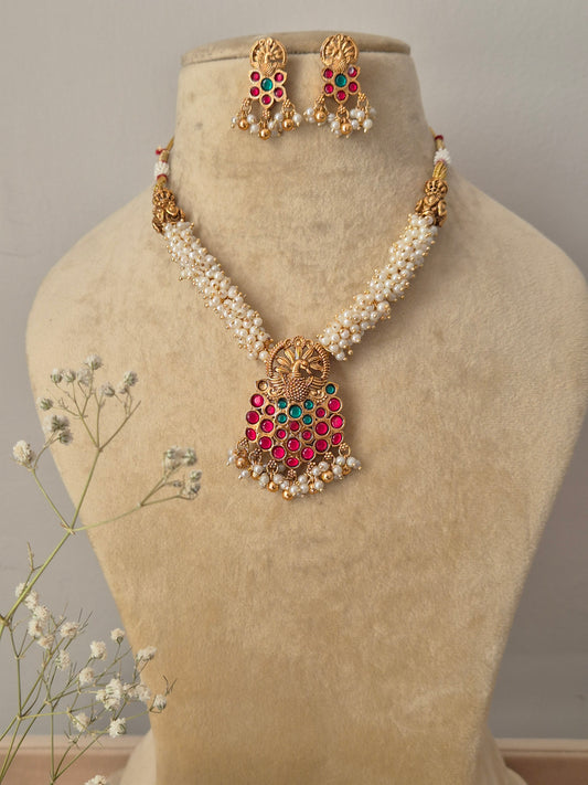 Handcrafted gold-plated temple jewelry pendant featuring a central deity motif, red and green stone accents, and pearl fringes on a multi-strand white pearl chain with matching stud earrings by naitri.in