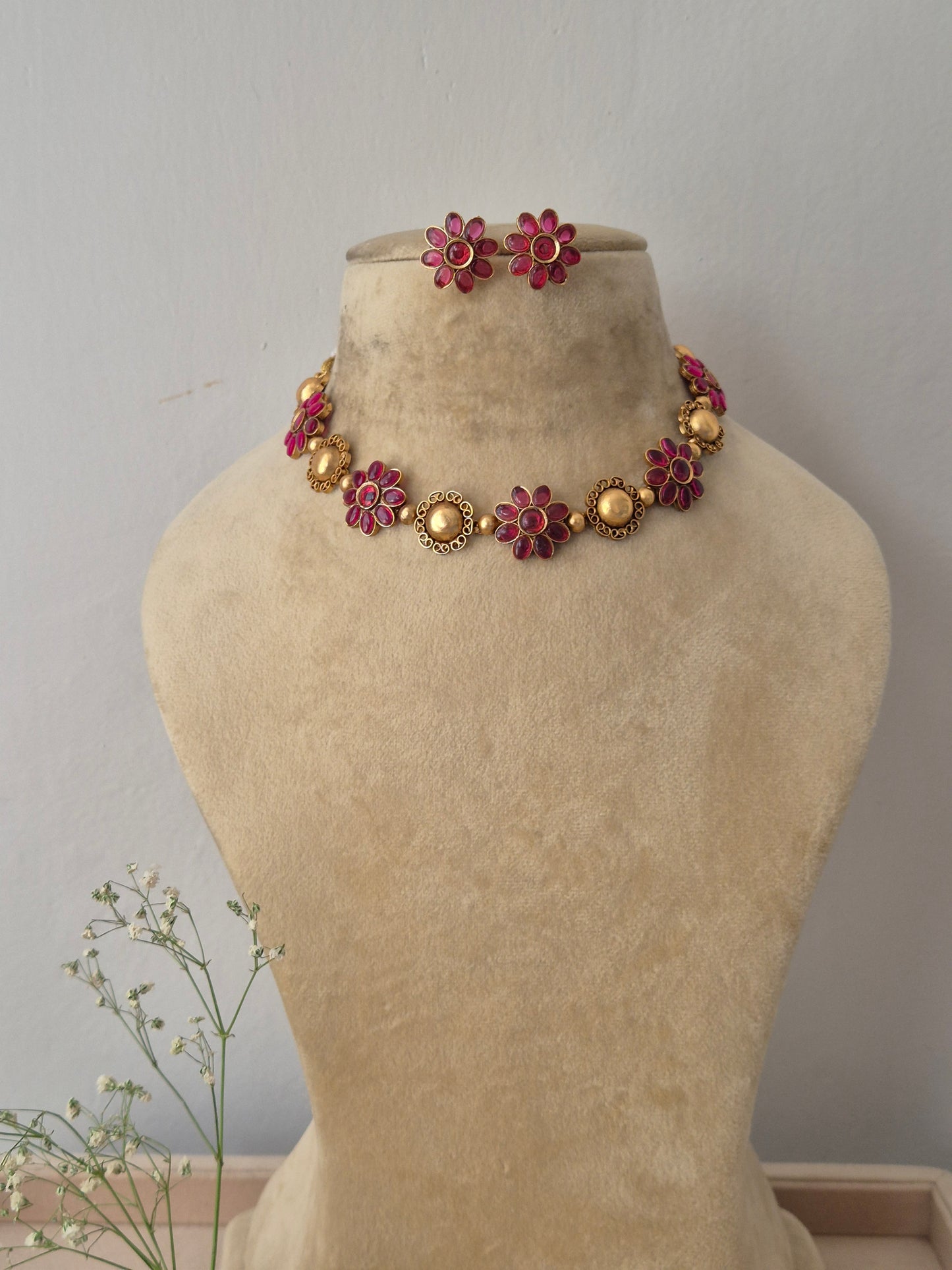 A gold-plated ethnic choker necklace featuring alternating textured round gold charms and red gemstone floral motifs,with matching flower stud earrings by naitri.in