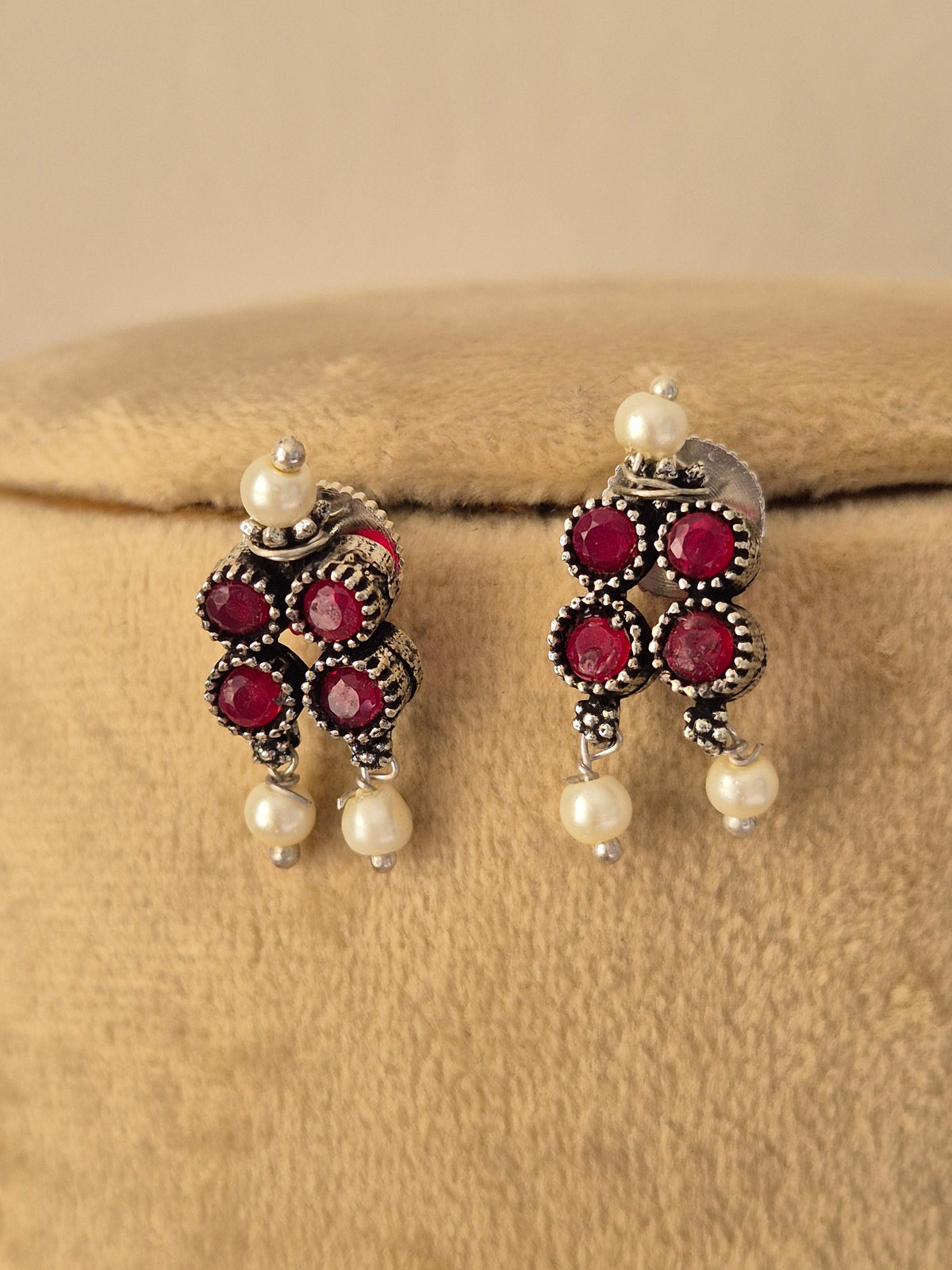 A pair of traditional Indian jhumka earrings with silver-toned metalwork, deep red round stones, and white pearl beads, by naitri.in