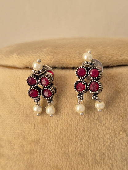 A pair of traditional Indian jhumka earrings with silver-toned metalwork, deep red round stones, and white pearl beads, by naitri.in