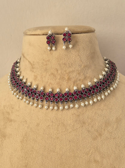 A traditional Indian jewelry set featuring a choker necklace and matching earrings, The set features silver-plated metal work, deep red stones, and white faux pearls by naitri.in