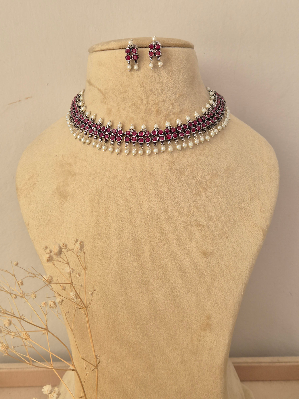 A traditional Indian jewelry set featuring a choker necklace and matching earrings the jewelry features silver-toned metal work, deep pinkish-red stones, and white faux pearls by naitri.in