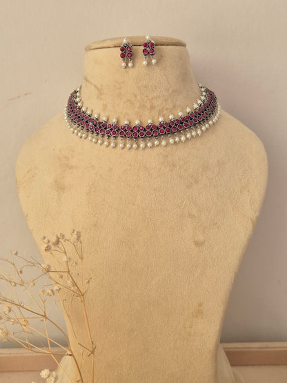 A traditional Indian jewelry set featuring a choker necklace and matching earrings the jewelry features silver-toned metal work, deep pinkish-red stones, and white faux pearls by naitri.in