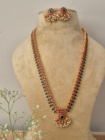 A long gold-plated traditional necklace featuring alternating red and green gemstone drops, a detailed central circular pendant, and hanging pearl clusters, by naitri.in