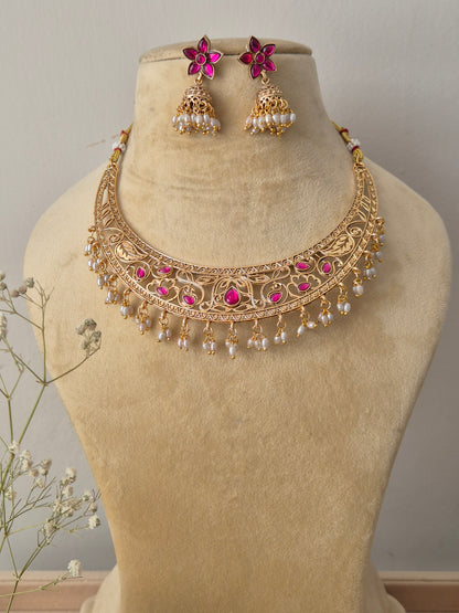 Handcrafted gold-plated temple jewelry choker necklace featuring intricate metal carvings, central ruby-pink stone accents, and hanging white pearl clusters, paired with matching pink floral jhumka earrings by naitri.in