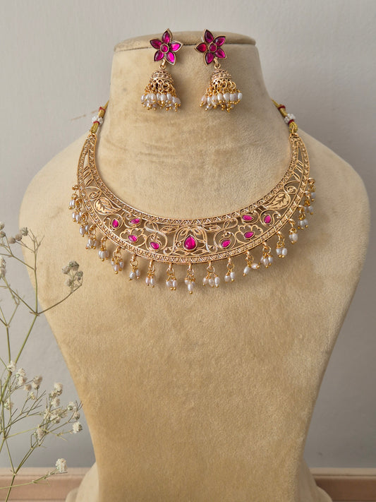 Handcrafted gold-plated temple jewelry choker necklace featuring intricate metal carvings, central ruby-pink stone accents, and hanging white pearl clusters, paired with matching pink floral jhumka earrings by naitri.in
