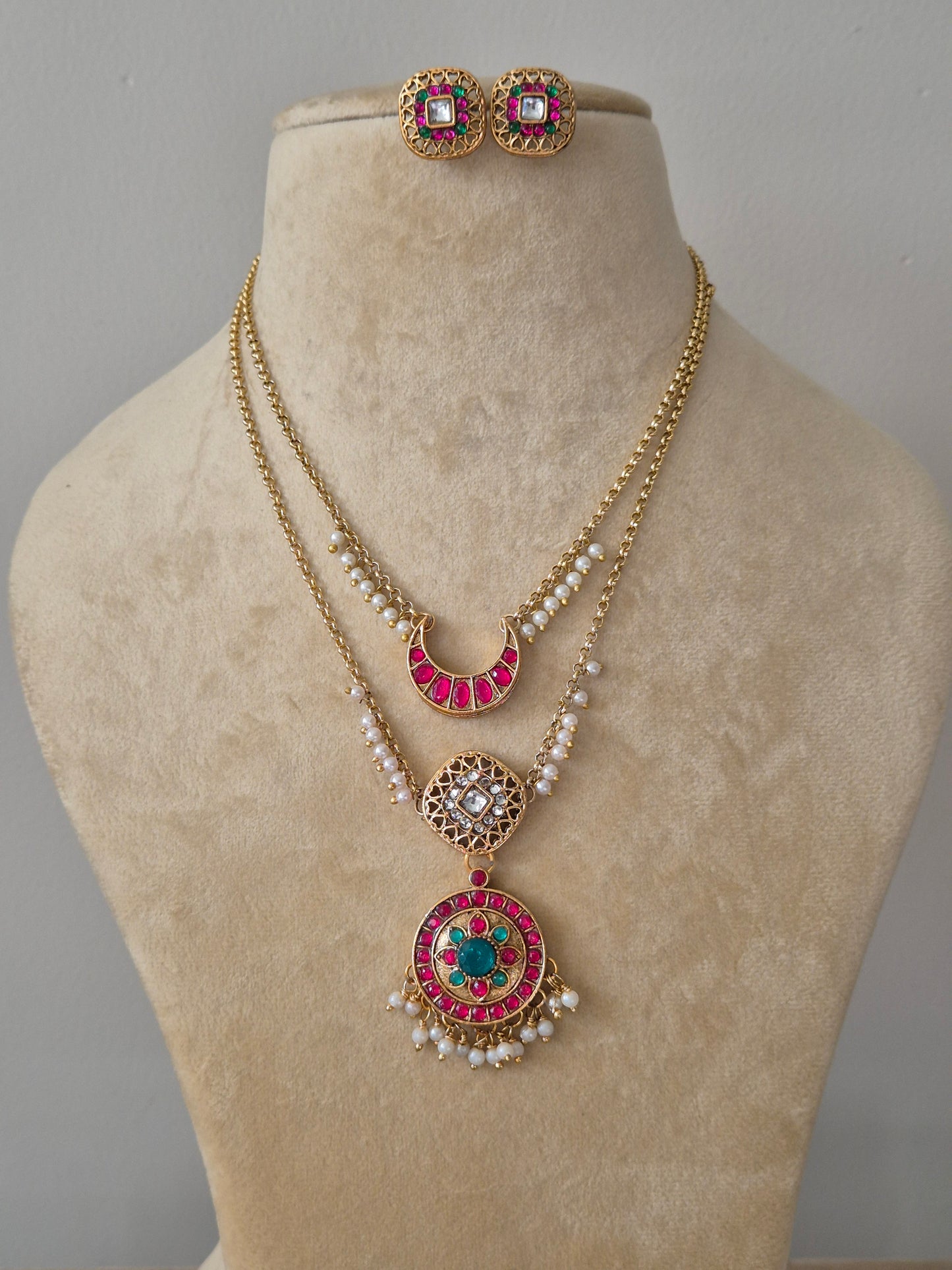 A gold-toned double-layered necklace set featuring a pink stone crescent and a circular pendant with green and pink accents,with matching square stud earrings By naitri.in