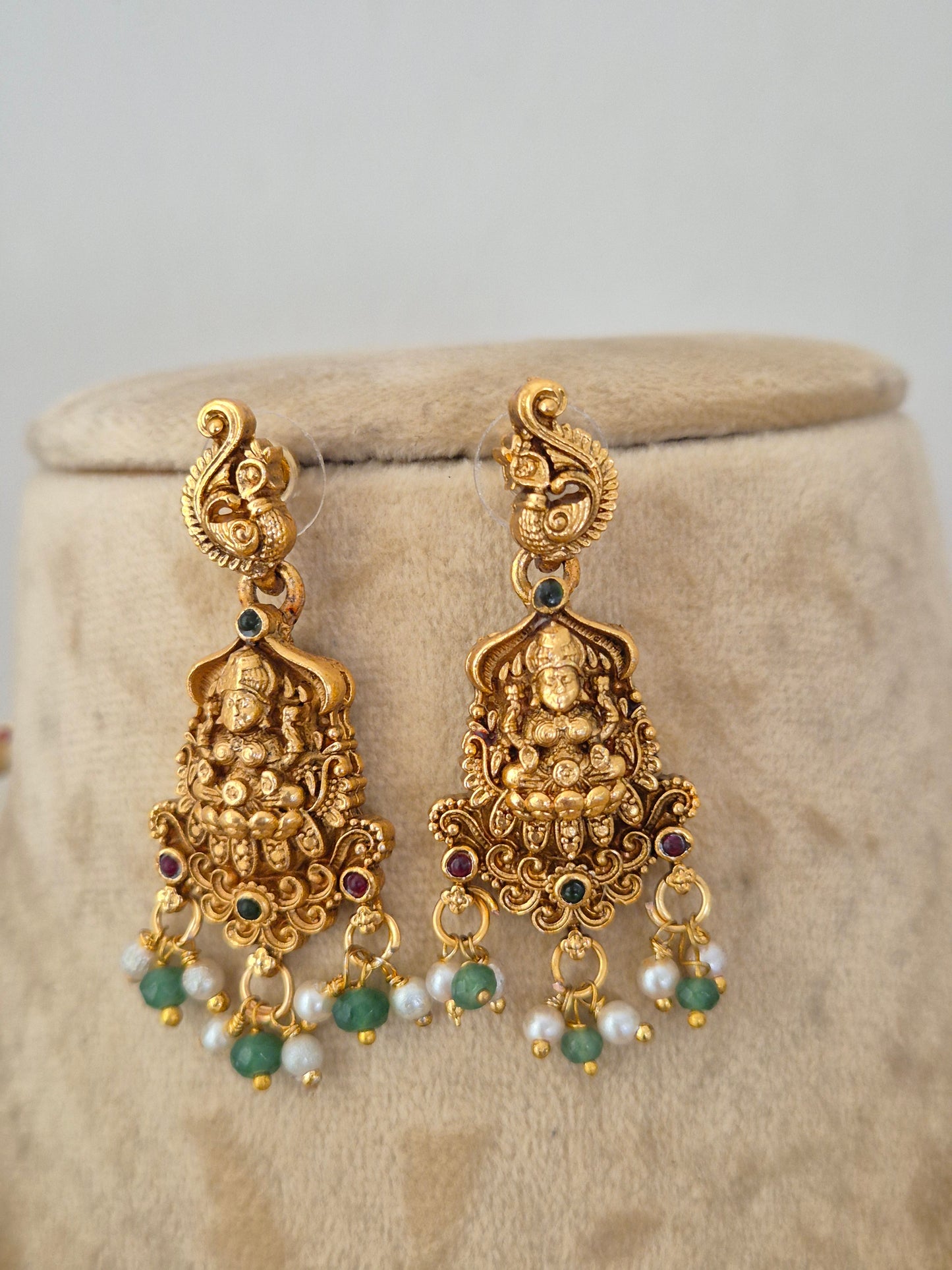 A pair of ornate gold-toned Indian jhumka earrings with detailed Ganesha motifs, small green stone accents, and white pearl beads, by naitri.in