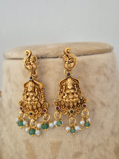A pair of ornate gold-toned Indian jhumka earrings with detailed Ganesha motifs, small green stone accents, and white pearl beads, by naitri.in