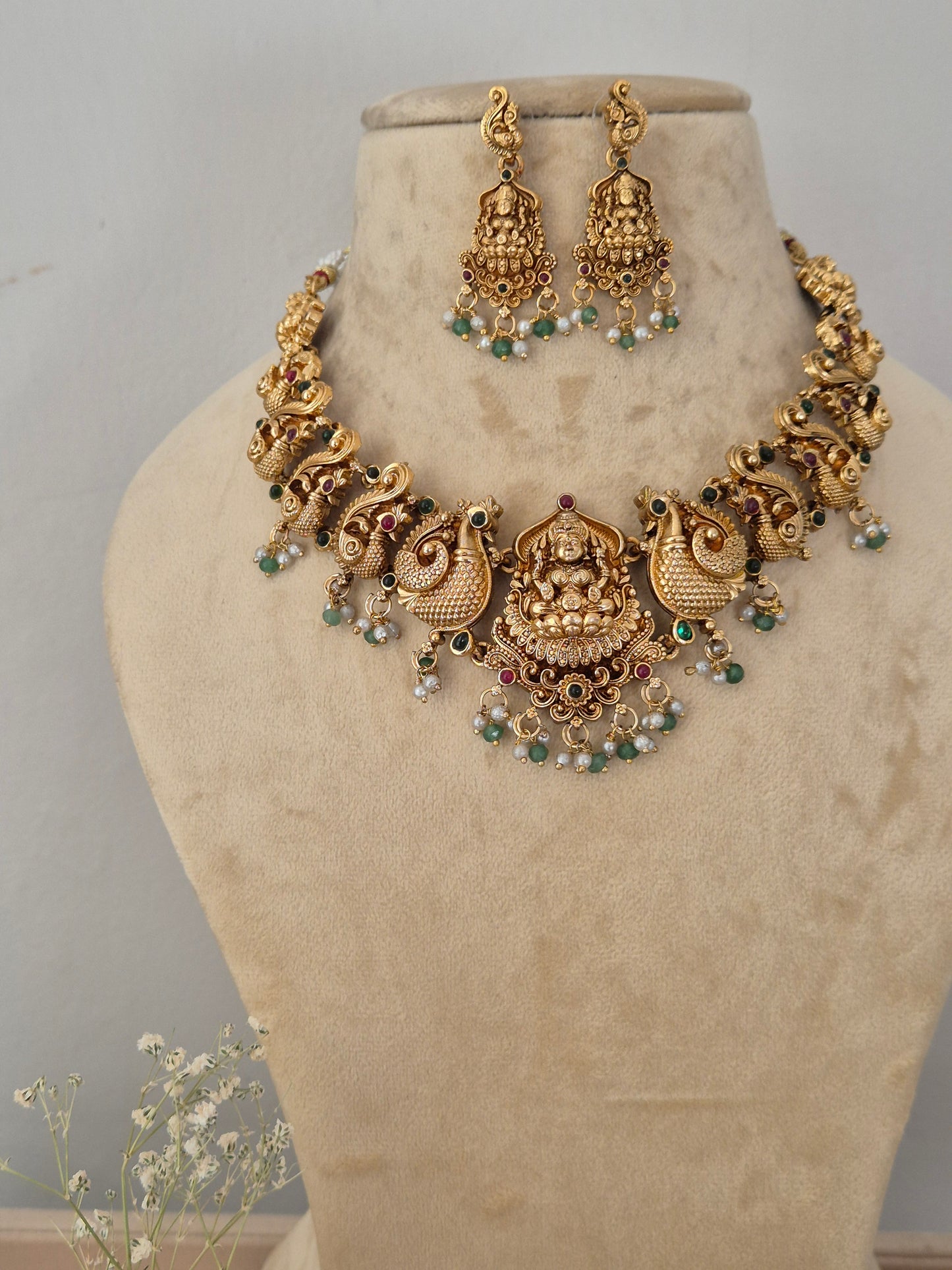 Antique gold-toned traditional Indian choker necklace and matching dangle earrings, the jewelry features intricate temple-style filigree work, engravings of deities, and a delicate fringe of hanging white faux pearls and small green beads by naitri.in