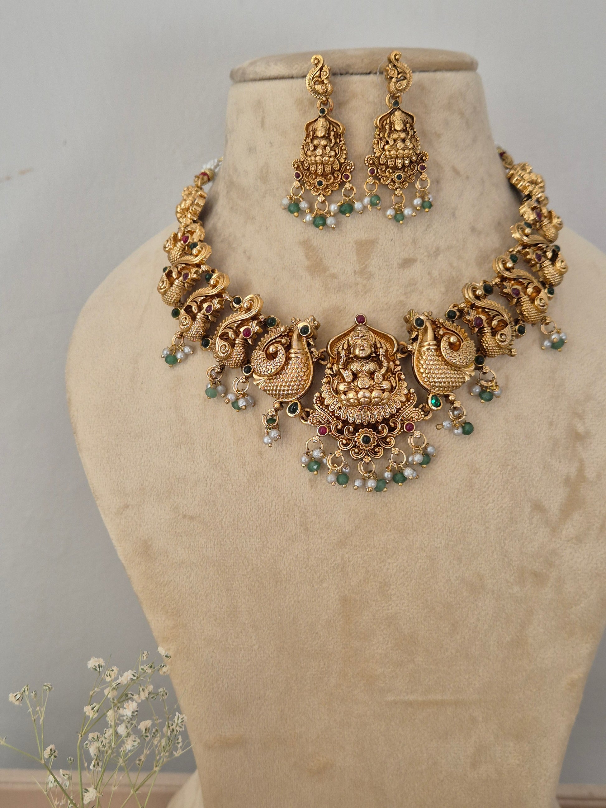 Antique gold-toned traditional Indian choker necklace and matching dangle earrings, the jewelry features intricate temple-style filigree work, engravings of deities, and a delicate fringe of hanging white faux pearls and small green beads by naitri.in