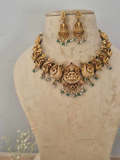 Antique gold-toned traditional Indian choker necklace and matching dangle earrings, the jewelry features intricate temple-style filigree work, engravings of deities, and a delicate fringe of hanging white faux pearls and small green beads by naitri.in