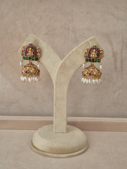 Pair of gold-plated Lochana Jhumka earrings hanging on a beige display stand, featuring rectangular temple-style studs with a Goddess motif, red and green gemstone accents, and bell-shaped drops fringed with small white pearls by naitri.in