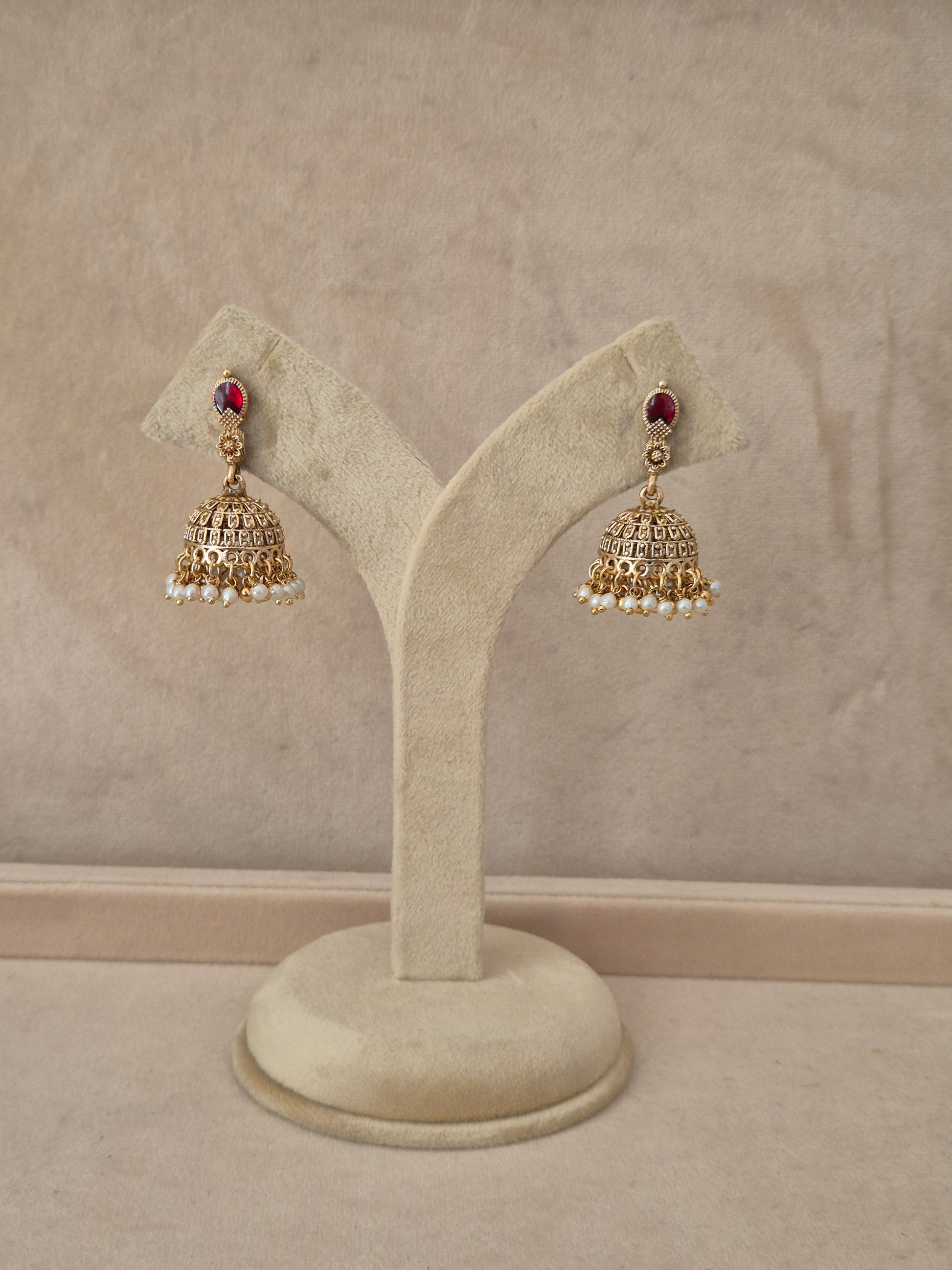 air of gold-toned Madhumita Jhumka earrings hanging on a beige velvet display stand, featuring teardrop-shaped studs with bright red gemstones and detailed bell-shaped drops fringed with small white pearls by naitri.in